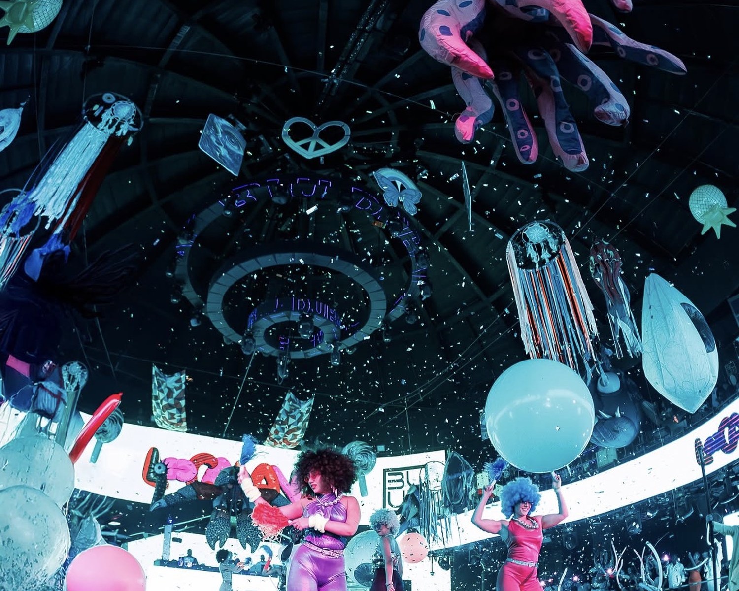 Costumed carnival performers on stage with giant balloons and confetti at a themed live event